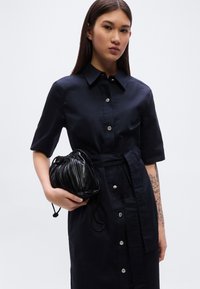 Φόρεμα μπλε ναυτικού με κουμπιά, κοντά μανίκια, σφιχτή μέση και απαλό ύφασμα. Συνδυάζεται με μαύρη τσάντα από δέρμα με πτυχώσεις.