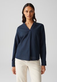 Camicia a maniche lunghe blu navy con scollatura a V, caratterizzata da un drappeggio morbido e polsini con bottoni, abbinata a pantaloni beige chiaro.