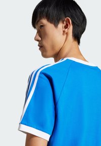 Blue athletic shirt with white trim and three white stripes on sleeves, featuring a crew neck and a smooth, lightweight fabric texture.