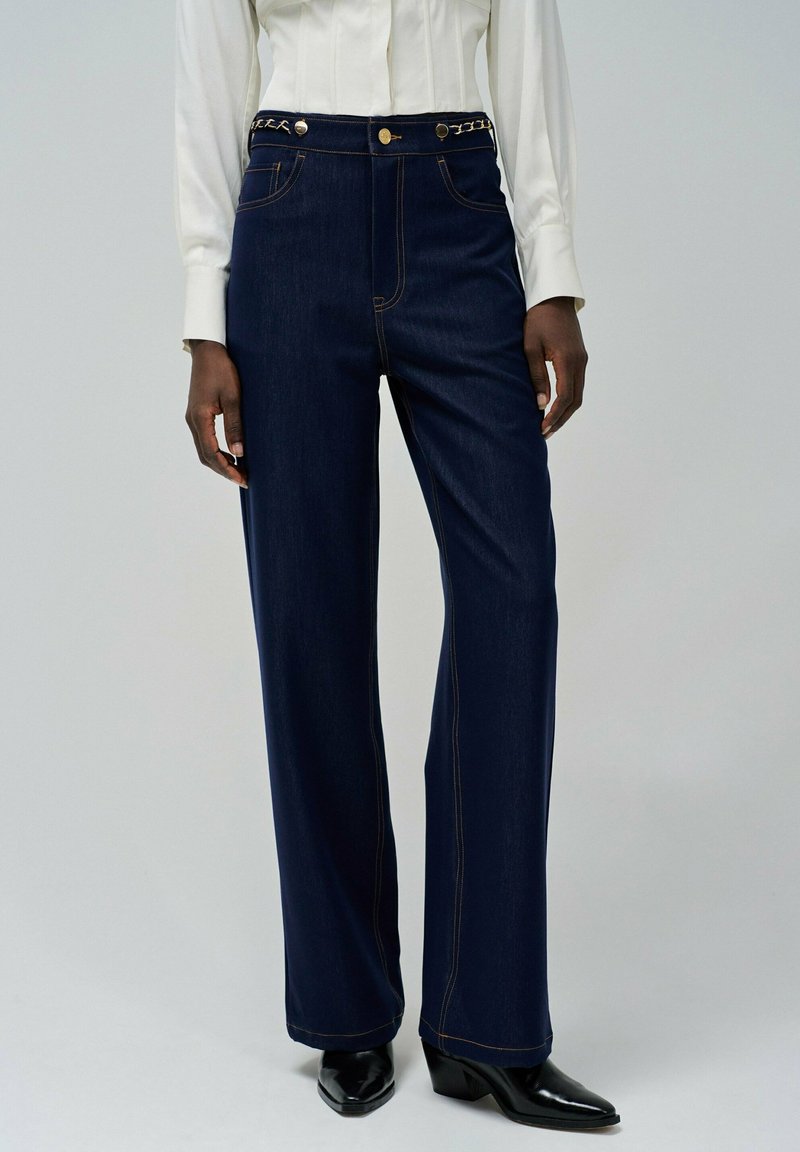 Jeans de tiro alto de color azul oscuro con costuras doradas y un detalle de cadena trenzada en la cintura, combinados con una camisa blanca ajustada y zapatos negros.