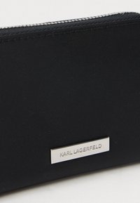 Melnā ar rāvējslēdzēju somiņa ar sudraba metāla logotipa plāksni, uz kuras ir izkrāsots "KARL LAGERFELD" uz auduma virsmas.