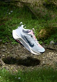 Chaussure de course Nike en gris et rose, présentant une surface texturée, des accents en mesh et un logo noir, avec une semelle intermédiaire blanche mouchetée.