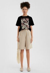 Zwarte t-shirt met bloemenprint en tekst, beige linnen shorts, tas van natuurlijke vezels. Instappers met een geweven patroon. Casual zomeroutfit.