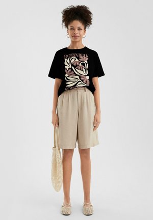 Zwarte t-shirt met bloemenprint en tekst, beige linnen shorts, tas van natuurlijke vezels. Instappers met een geweven patroon. Casual zomeroutfit.