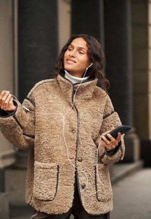Femme portant un manteau marron duveteux, tenant un téléphone et ajustant des écouteurs filaires, debout à l'extérieur près d'un bâtiment avec des colonnes.