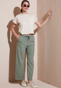 Cremfarbendes, kurzes T-Shirt mit Spitzen-Pocket-Detail, kombiniert mit hellgrünen, weit geschnittenen Hosen und weißen Sneakers vor einem strukturierten Hintergrund.