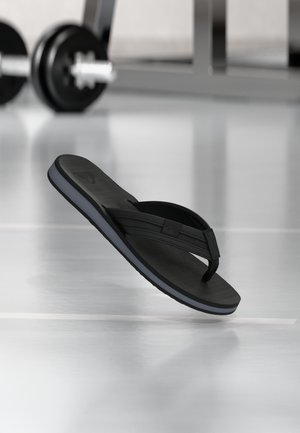 Claquette noire flottant au-dessus d'un sol de gym scintillant avec une barre de musculation floue en arrière-plan.