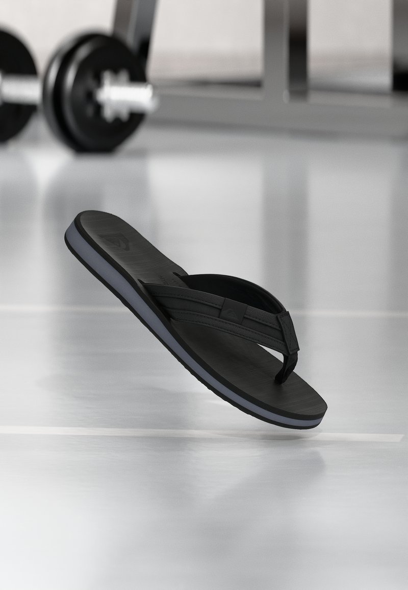 Fekete papucs lebeg egy fényes edzőtermi padló felett, háttérben elmosódott súlyemelő rúddal.