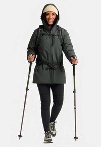 Dark green waterproof jacket with drawstring, black leggings, beige beanie, and hiking poles. Features a zippered front and adjustable cuffs.