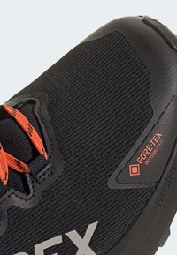 La chaussure de sport noire présente une tige en tissu texturé, des lacets orange et un logo GORE-TEX bien visible sur le côté. Semelle en caoutchouc épaisse pour une meilleure adhérence.