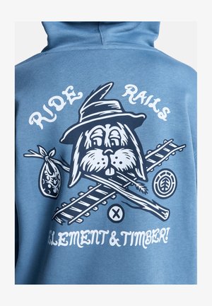 Blaue Kapuzenjacke mit einem Grafikdesign eines Kaninchens mit Hut, gekreuzten Ästen und einer Tasche. Der Text lautet "RIDE RAILS ELEMENT & TIMBER!" in Weiß.