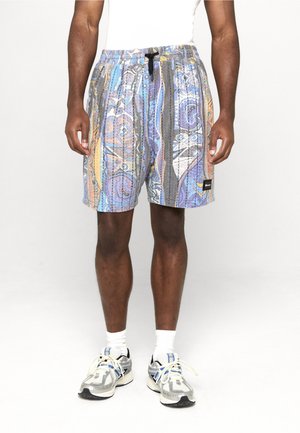 Hombre con pantalones cortos de patrón colorido, calcetines blancos y zapatos deportivos grises, de pie contra un fondo claro y liso.