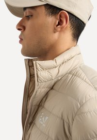 Giacca puffer beige con colletto alto, texture trapuntata, zip frontale e logo riflettente. Accompagnata da un cappellino neutro.
