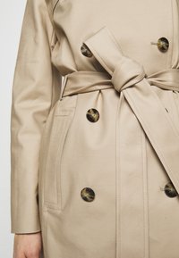 Trench-coat beige avec des boutons en écaille foncée, ceinture nouée à la taille, poches avant et manches longues sur le torse d'une personne.