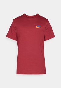 T-shirt rouge à manches courtes et col rond avec un petit logo Nike bleu et orange sur le côté gauche de la poitrine, sur un fond uni.