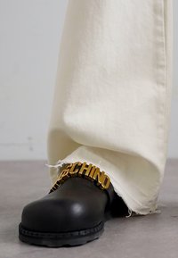 Μαύρο δερμάτινο παπούτσι με στρογγυλή μύτη και χρυσές λεπτομέρειες λογότυπου. Συνδυασμένο με λευκά παντελόνια με φθαρμένο στρίφωμα, τοποθετημένο σε γκρίζα επιφάνεια.