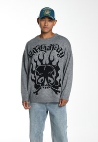Grauer Strickpullover mit einem großen schwarzen Totenkopf- und Flammenmotiv. Das Model trägt hellblaue Jeans.