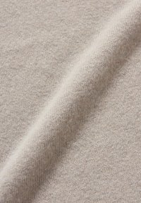 Helles, beiges Gewebe mit einer weichen, strukturierten Oberfläche, das ein dezentes Strickmuster und einen leichten Glanz aufweist. Das Material wirkt flexibel und warm.