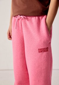 Person wearing pink textured sweatpants with drawstring and "AMERICAN VINTAGE" logo on thigh, hands in pockets, brown top visible.