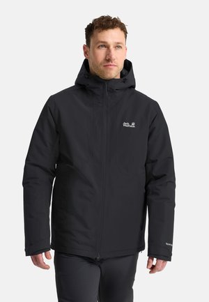 Man draagt een zwarte Jack Wolfskin capuchonjas en zwarte broek, staand en licht naar rechts kijkend op een witte achtergrond.