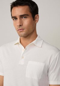 Bílá polo košile vyrobená z měkké látky s klasickým límečkem, třemi knoflíky a kapsou na levé straně hrudi. Má čistý, minimalistický design.
