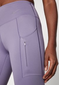 Lila sportos leggings áramvonalas kialakítással, zippel zárható zsebbel és diszkrét varrásokkal a cipzáros megjelenésért és sima textúráért.