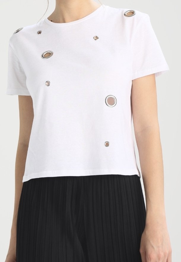 Crop top blanc à manches courtes avec découpes à œillets métalliques, associé à une jupe plissée noire ou un pantalon, porté par une personne sur un fond uni.