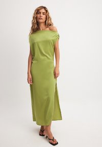 Off-shoulder, ilgis iki žemės suknelė iš ryškiai žalio satino. Turi šoną plyšį ir minkštą, lygų tekstūrą. Atviros rankovės, suteikiančios laisvą palaidinimą.