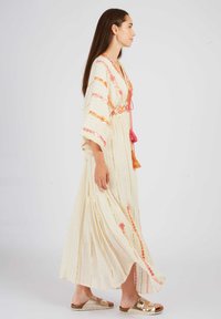 Langes, cremefarbenes Maxikleid mit floralen Stickereien in Pink und Orange, mit einer Schleife in der Taille und weiten Ärmeln. Kombiniert mit Sandalen.