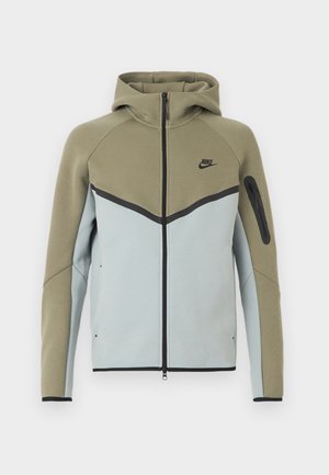 Zipējama Nike džemperis ar olīvu zaļu augšdaļu, gaiši pelēku apakšu, melnu apmalējumu, piedurknes kabatu ar rāvējslēdzēju un piešūtu kapuci, attēlots no priekšpuses.