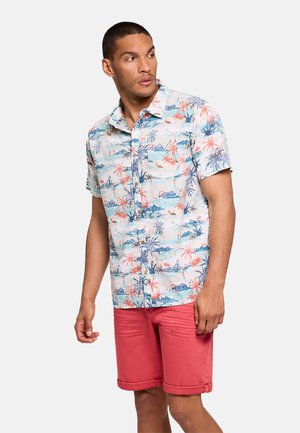 Hombre con camisa de manga corta abotonada con estampado tropical y pantalones cortos de color coral, mirando hacia su izquierda contra un fondo liso.