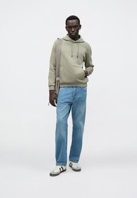 Khaki hettegenser med snorer, frontlomme; lyseblå jeans med rette ben; grå og hvite joggesko med svarte striper; bærer en skulderveske.