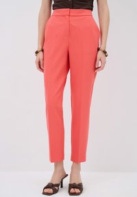 Coral tailored trousers with a high waist, slim fit, and crease detail. Worn with brown heeled sandals, showcasing smooth fabric and clean lines.
