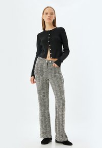 Svart ribbad cardigan med knappstängning, tillsammans med strukturerade svart- och vitrandiga utsvängda byxor. Modellen står med ena handen i fickan.