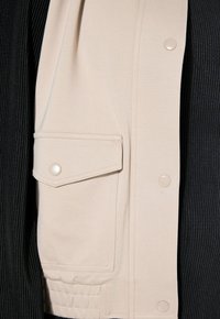 Beżowa bezrękawnik z dużą boczną kieszenią, zatrzaskami i miękką fakturą, zestawiona z czarnym, prążkowanym topem pod spodem.