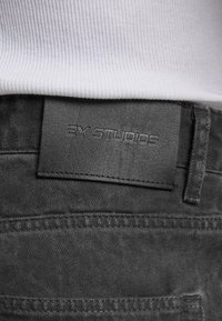 Černá kožená nášivka s embosovaným nápisem "2Y STUDIOS" na tmavě šedých džínech, které jsou nošeny s bílým žebrovaným topem.