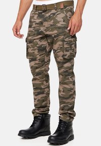 Vihreän ja ruskean sävyiset maastokuvioidut cargo-housut, joissa on useita taskuja ja vyö, yhdistettynä mustiin nauhallisiin saappaisiin.