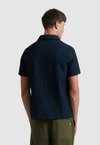 Chemise à manches courtes, bleu marine, avec un motif texturé, dotée d'un col et d'une coupe décontractée, associée à un pantalon vert olive.