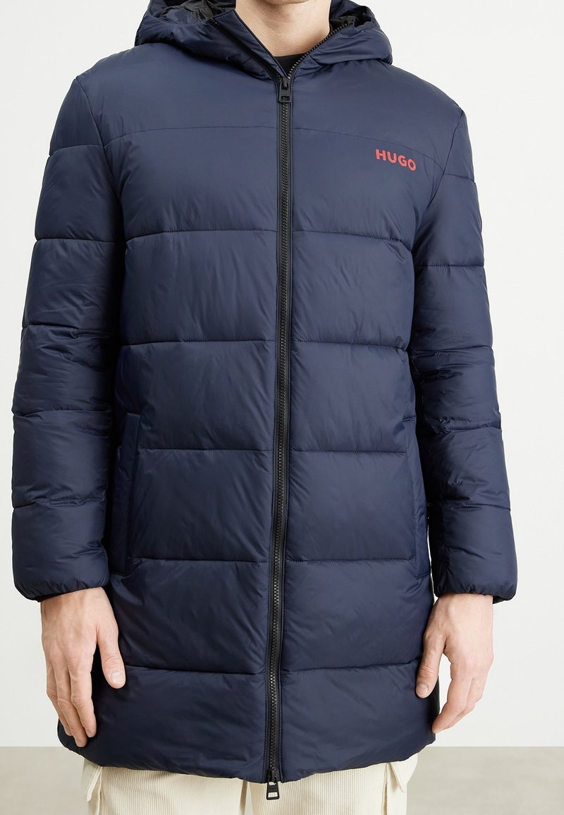 Chaqueta de plumas azul marino con paneles acolchados horizontales, cierre frontal con cremallera y capucha. El logo de la marca "HUGO" se muestra en rojo.