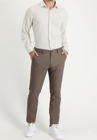 Man wearing a light beige long-sleeve button-up shirt, brown slim-fit trousers, white sneakers, standing with hands in pockets.