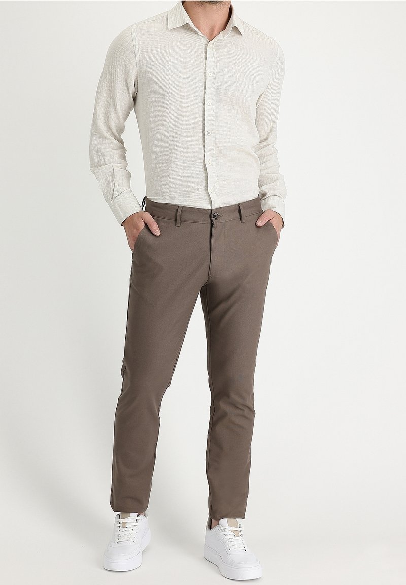 Man wearing a light beige long-sleeve button-up shirt, brown slim-fit trousers, white sneakers, standing with hands in pockets.