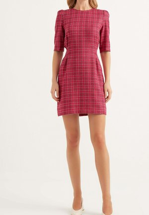 Mujer con un vestido ajustado de cuadros rojos, mangas hasta el codo y zapatos blancos de tacón alto, de pie frente a un fondo liso.