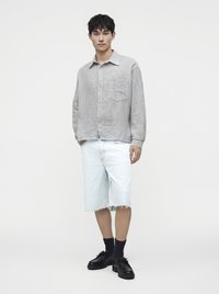 Jeune homme portant une chemise grise à manches longues, un short en jean clair, des chaussettes noires et des chaussures noires, debout devant un fond uni.