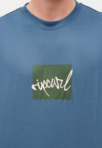 Μπλε κοντομάνικη μπλούζα με στρογγυλή λαιμόκοψη, με ένα πράσινο τετράγωνο και λευκή στιλιζαρισμένη επιγραφή "ripcurl" τυπωμένη στο στήθος.