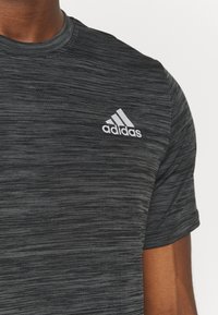 Lähikuva henkilöstä, joka pitää yllä tummanharmaata, kuvioitua Adidas-paitaa, jossa on Adidas-logo vasemmassa rinnassa.