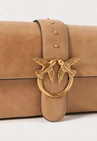 Tan zilo soma ar apaļo zelta aksesuāru, kas rotāts ar diviem putnu motīviem, izceltām ar zelta kniedēm uz siksnas un kontrastējošu stitchingu.