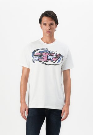 Ung mand iført en hvid t-shirt med et stort, abstrakt pink og blåt grafisk design på brystet, parret med mørke jeans.