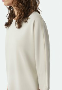Helle beige Oversized-Sweatshirt mit rundem Halsausschnitt, weicher Textur und kontrastierenden Nahtdetails entlang der Ärmel.