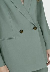 Dubbelgevoerde blazer in een gedempt groene stof, met twee grote donkere knopen, zijzakken en een zachte textuur. Eenvoudig, gestructureerd ontwerp.