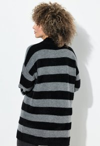 Vrouw met krullend haar die een losse zwart-grijze horizontaal gestreepte trui draagt, zijwaarts gekeerd tegen een witte achtergrond.